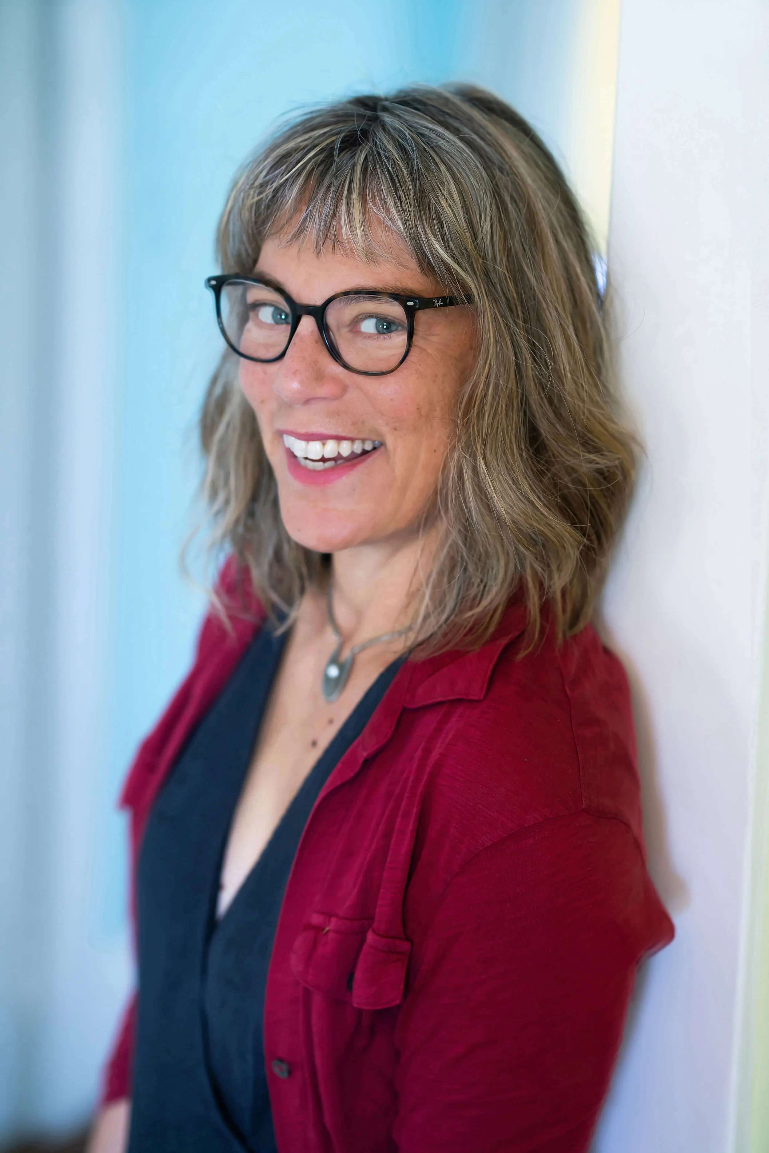 Dashka Slater smiling while leaning against a wall in a bright, softly lit setting.