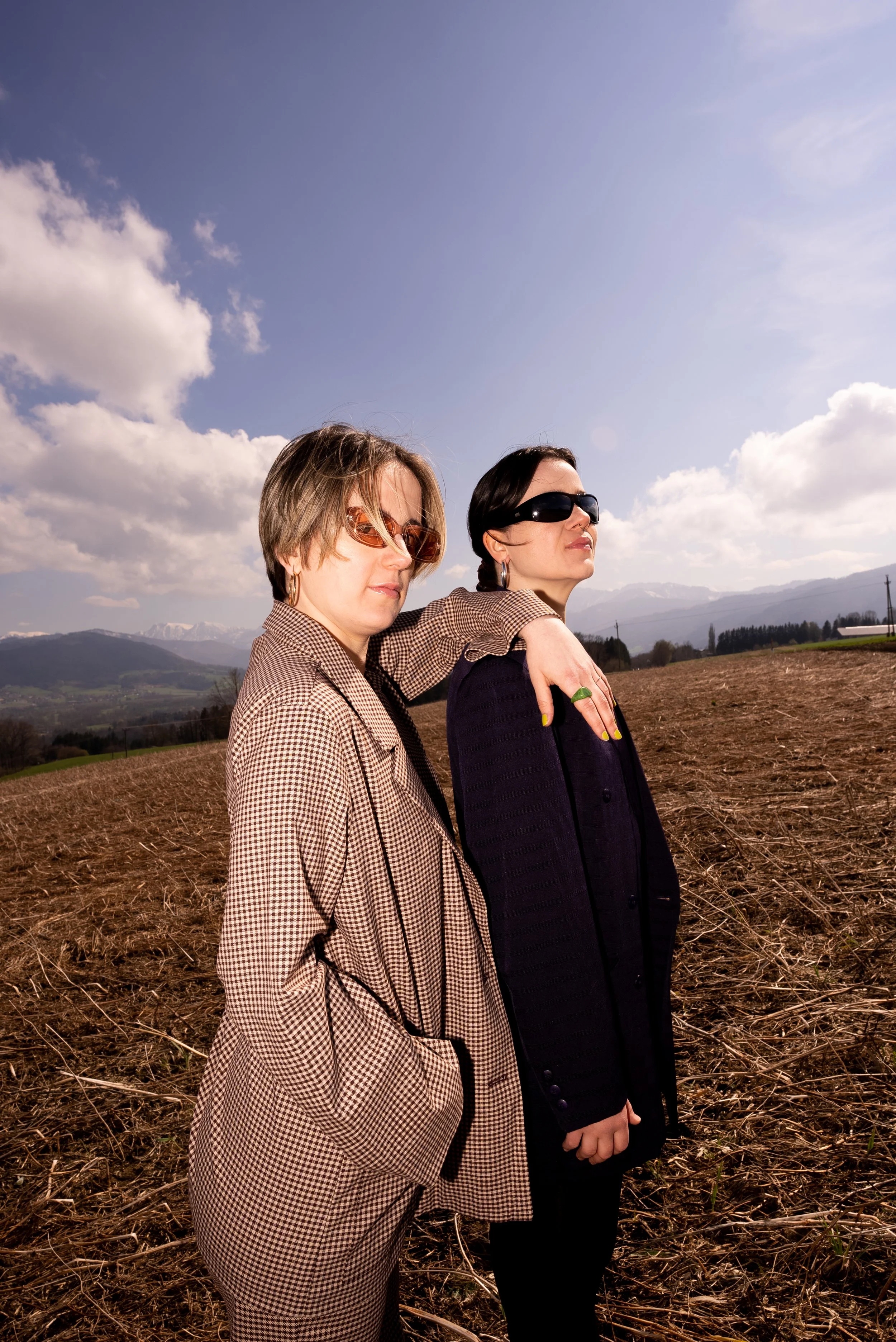 Zwei Frauen mit Sonnenbrillen stehen im Sonnenlicht auf einem Feld, umgeben von Bergen und Himmel mit Wolken.