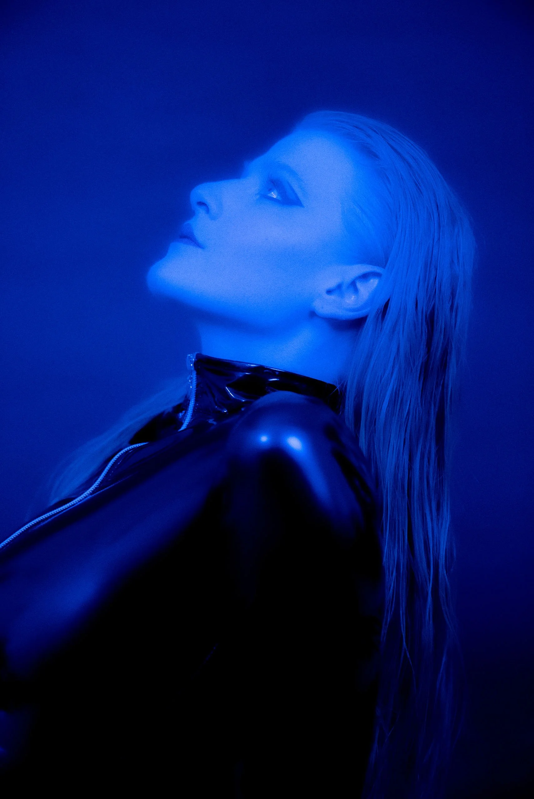 Porträt eines Models in Schwarzlicht mit blauer Beleuchtung, trägt eine schwarze, glänzende Jacke und hat lange, nasse Haare.