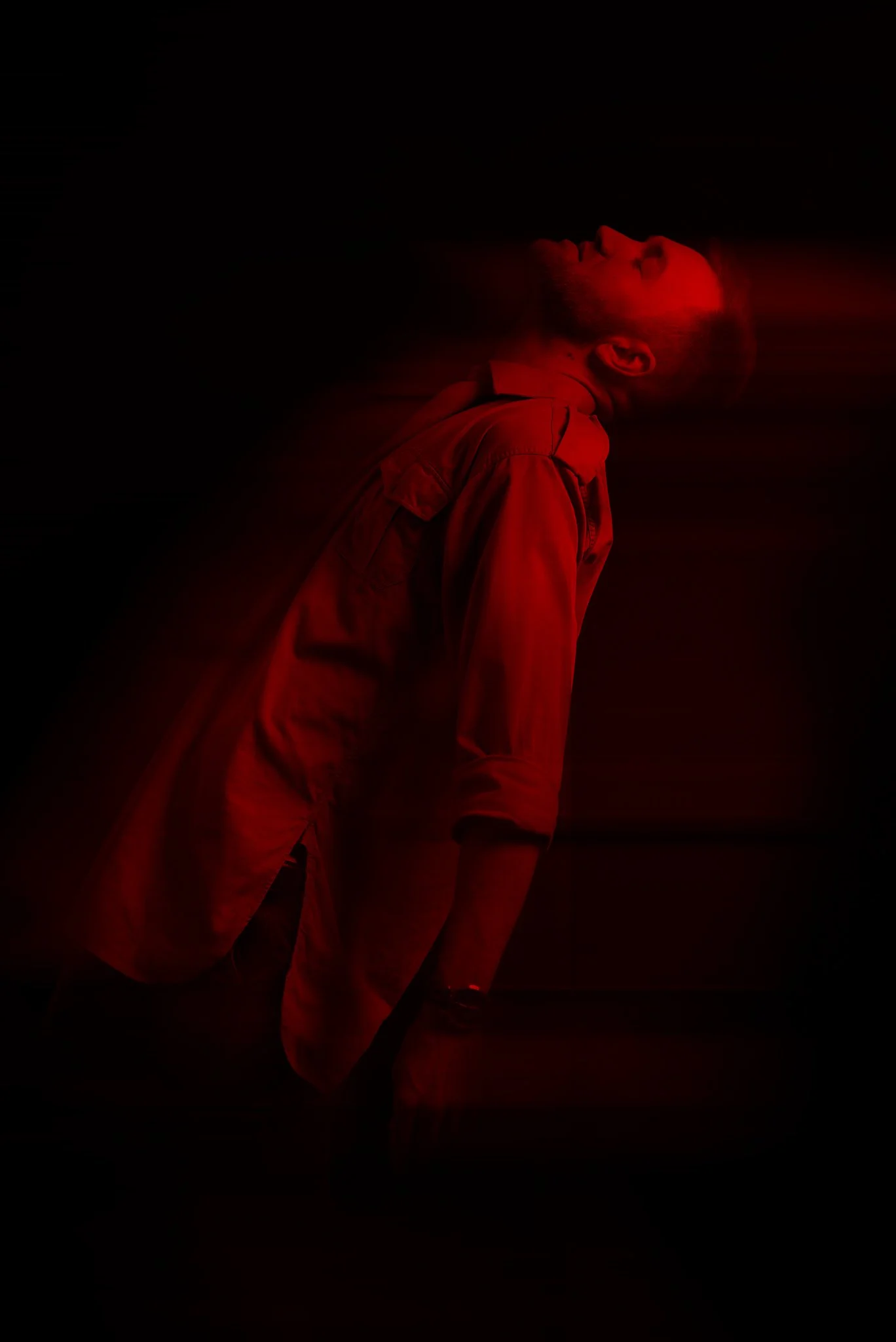 Ein Mann lehnt mit geschlossenen Augen in einem dunklen Raum, beleuchtet von rotem Licht, mit einer verschwommenen Bewegungseffekt.