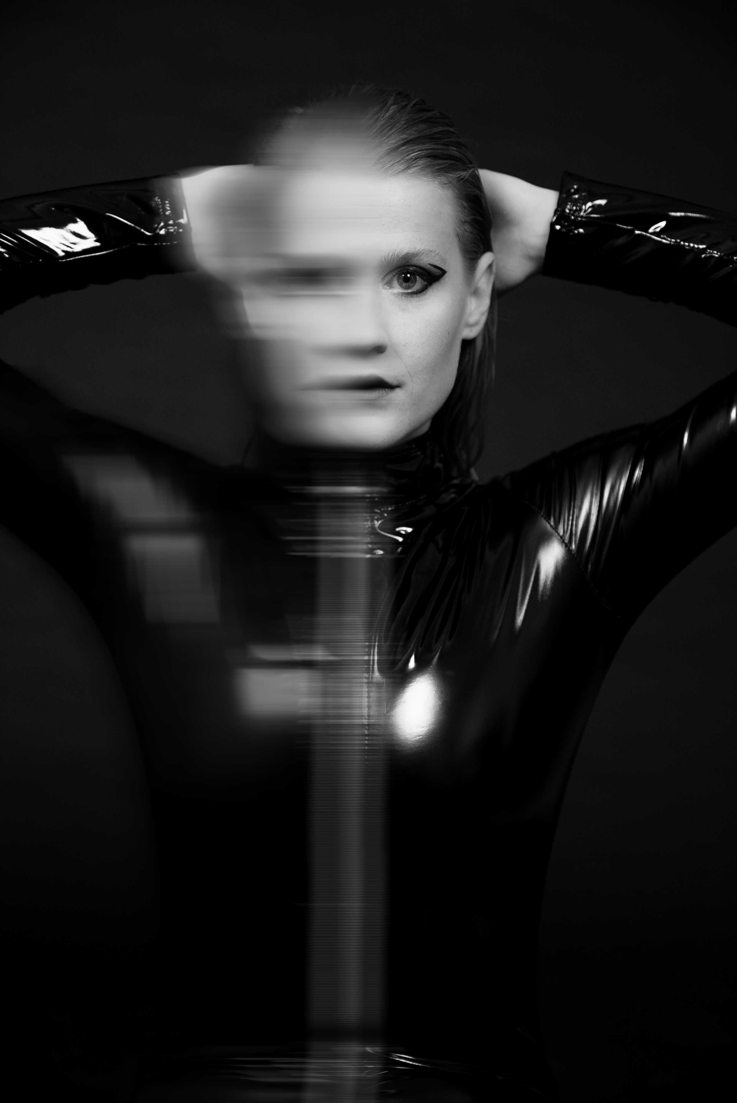 Schwarz-weiß-Fotografie einer Frau in einem glänzenden Lack-Outfit, die vor einem dunklen Hintergrund steht, mit halb verdecktem Gesicht, das nur eine Hälfte sichtbar ist und auf das andere schielt.