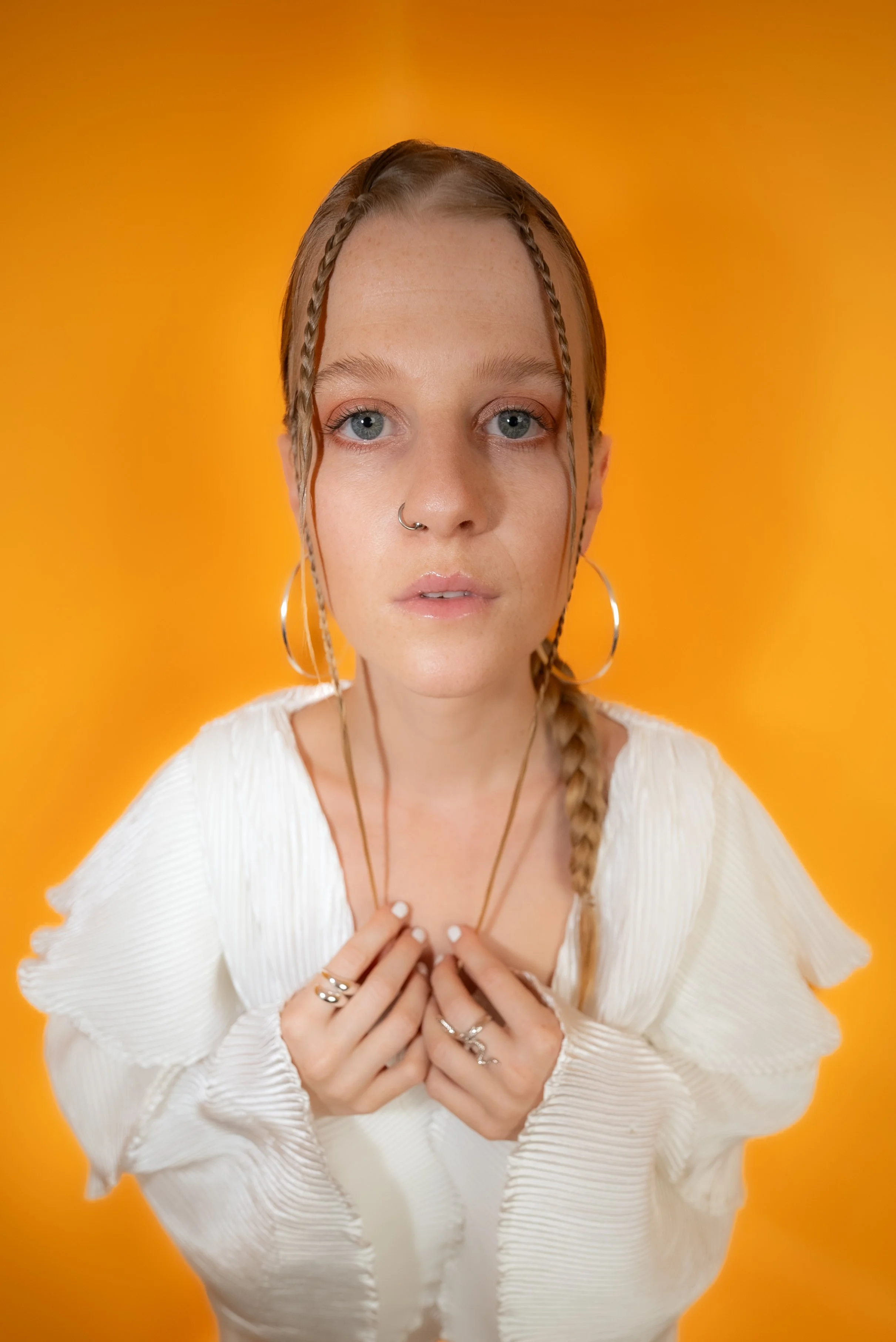 Porträt einer jungen Frau mit blonde Zöpfen vor orangem Hintergrund, trägt große Ohrringe, mehrere Ringe und einen weißen, gerüschten Pullover, schaut direkt in die Kamera.
