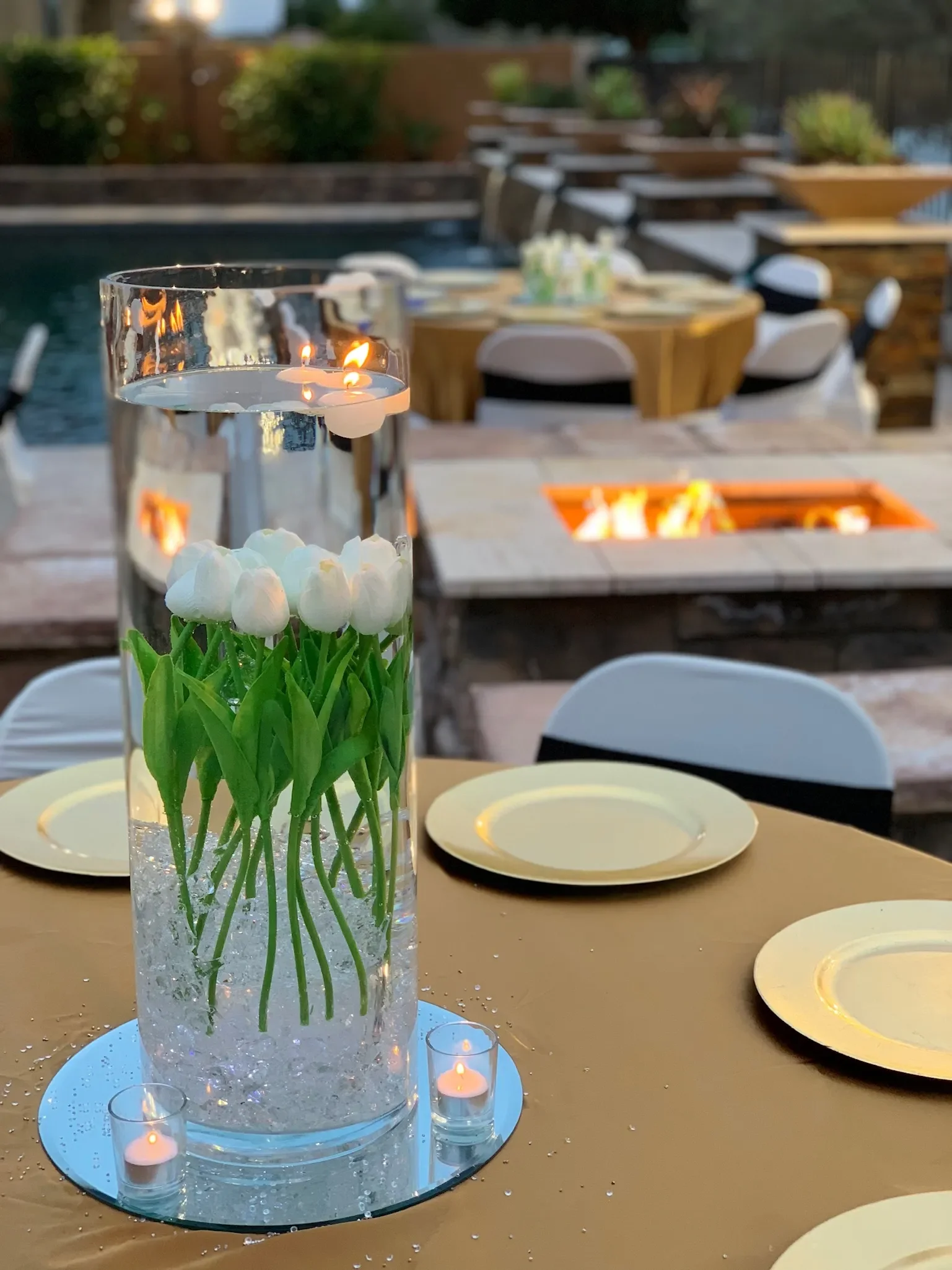 A tall glass vase with floating candles and white tulips serves as a centerpiece on an outdoor dining table. The table is set with yellow plates and surrounded by chairs, with a fire pit and additional tables in the background.