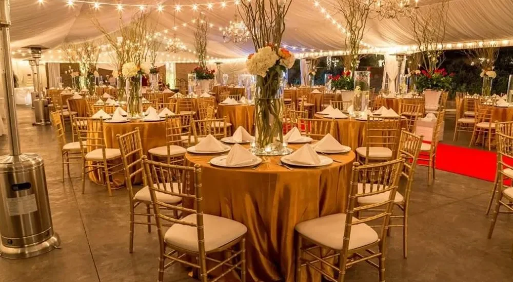 Elegant event tent decorated with fairy lights, tall floral centerpieces, and round tables with gold tablecloths and matching chairs.