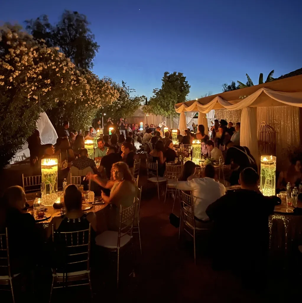 Outdoor evening event with people seated at decorated tables, illuminated by glowing lights, under a canopy and near flowering trees.