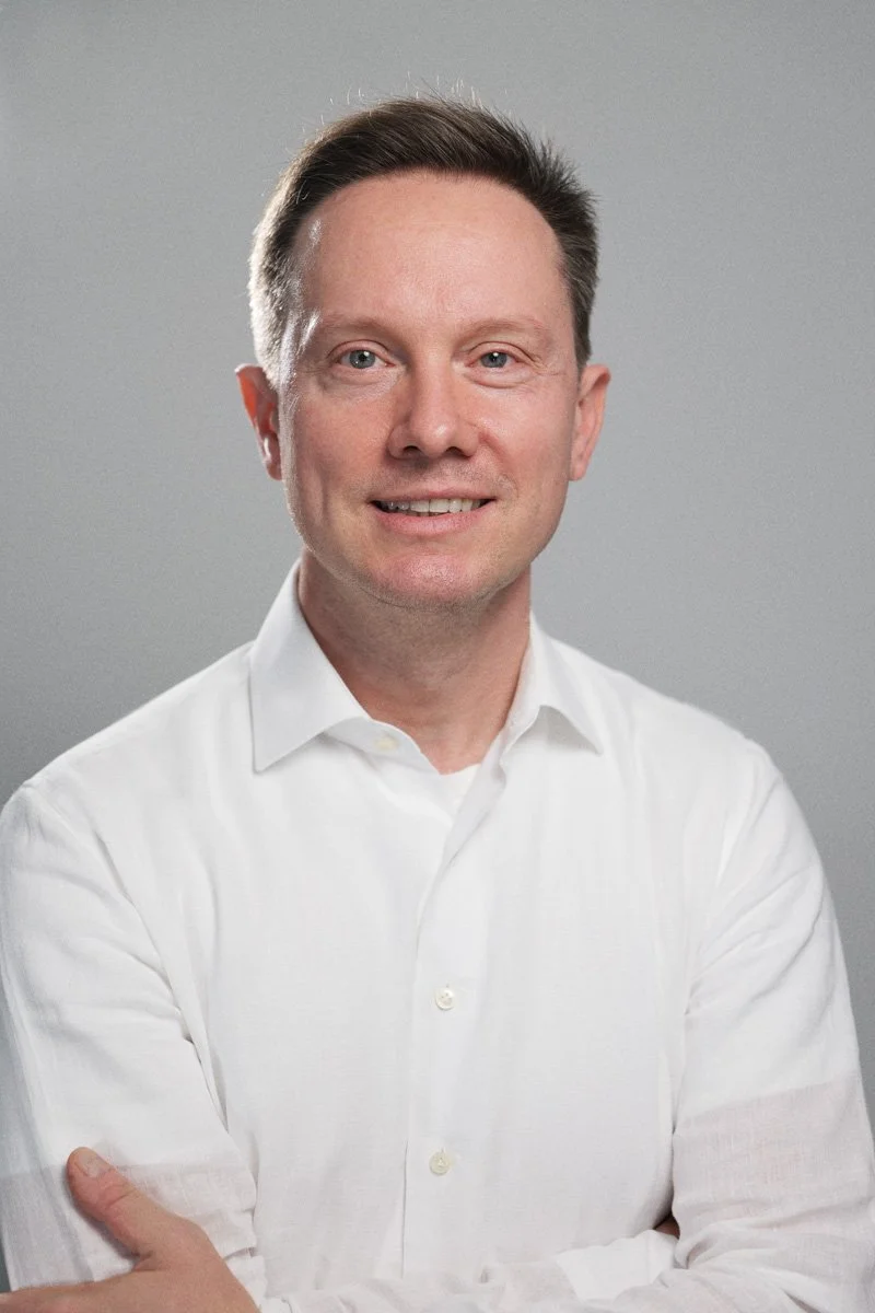 Porträt eines Mannes mit dunklem Haar, weißem Hemd, lächelnd vor grauem Hintergrund.