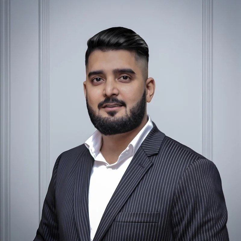 A man with dark, curly hair, wearing clear glasses and a nose ring, sitting indoors near a light-colored wall and a houseplant.