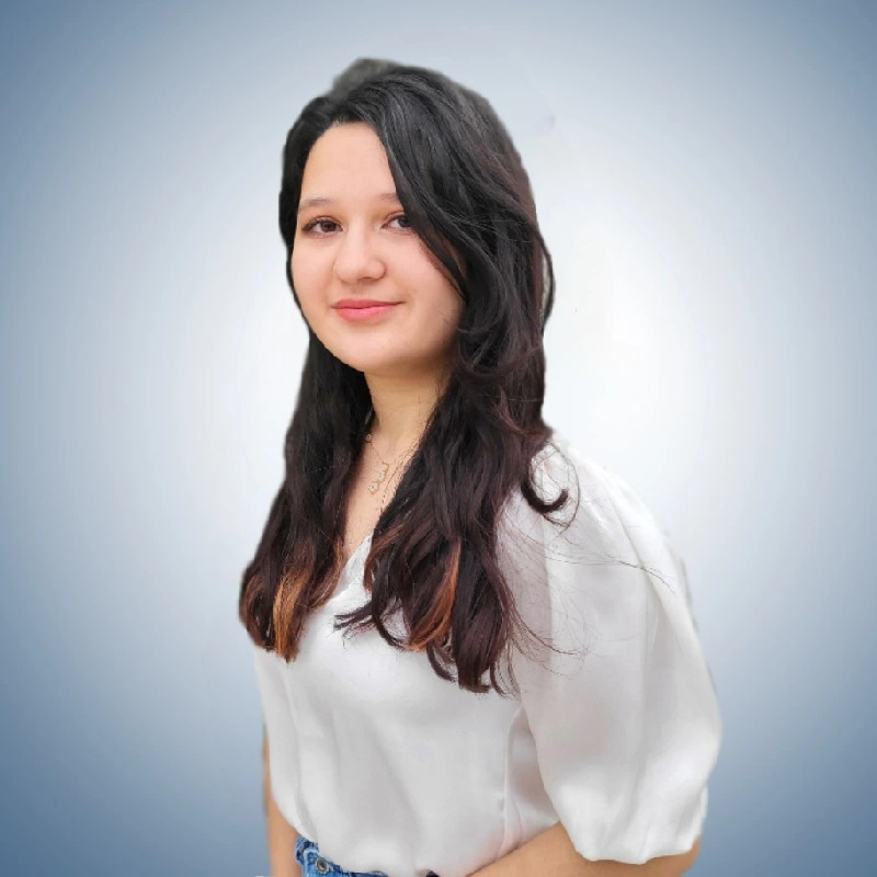 A man with curly dark hair, glasses, a nose ring, and a beard, wearing a dark sweater, looking to the side in a well-lit room.