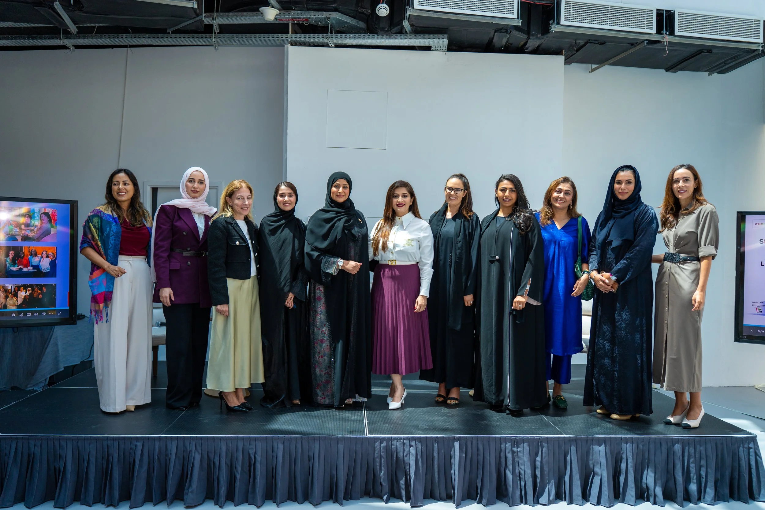 Group of diverse women standing on a stage at a formal event in an indoor venue, with two monitors on either side.