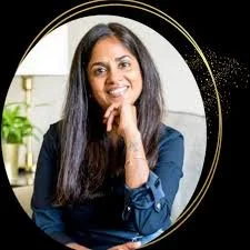 A woman with curly hair, glasses, and earrings smiling indoors with books and a window in the background.