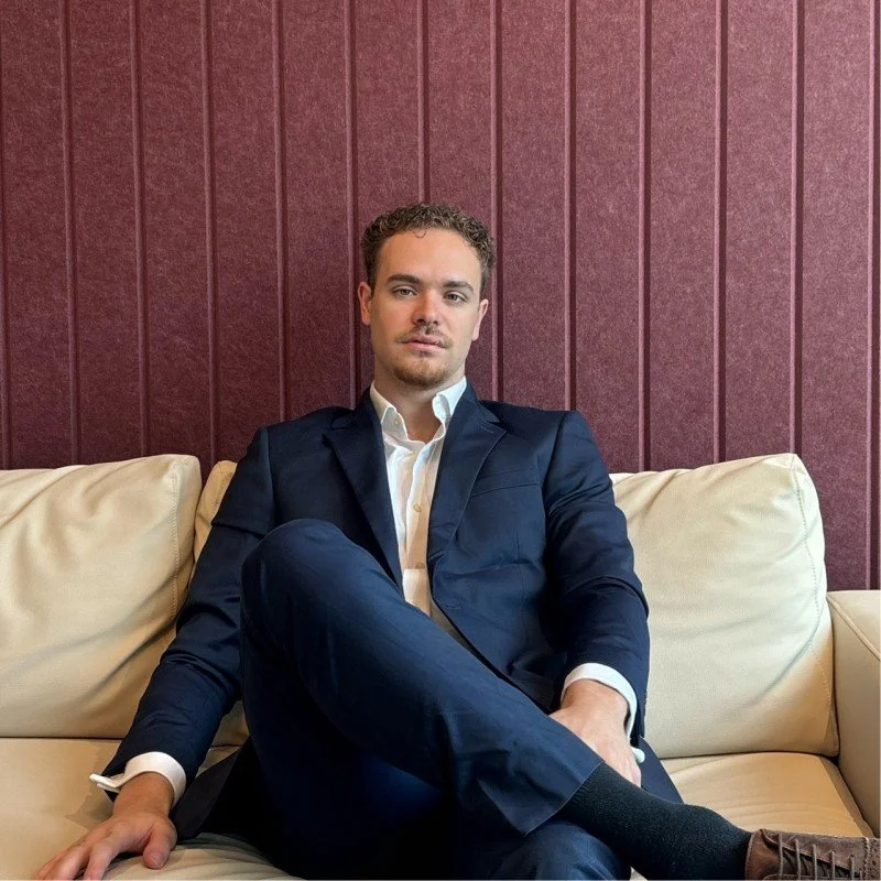 A man with curly dark hair, glasses, a nose ring, and a beard, wearing a dark sweater, looking to the right with a slight smile in a bright room.