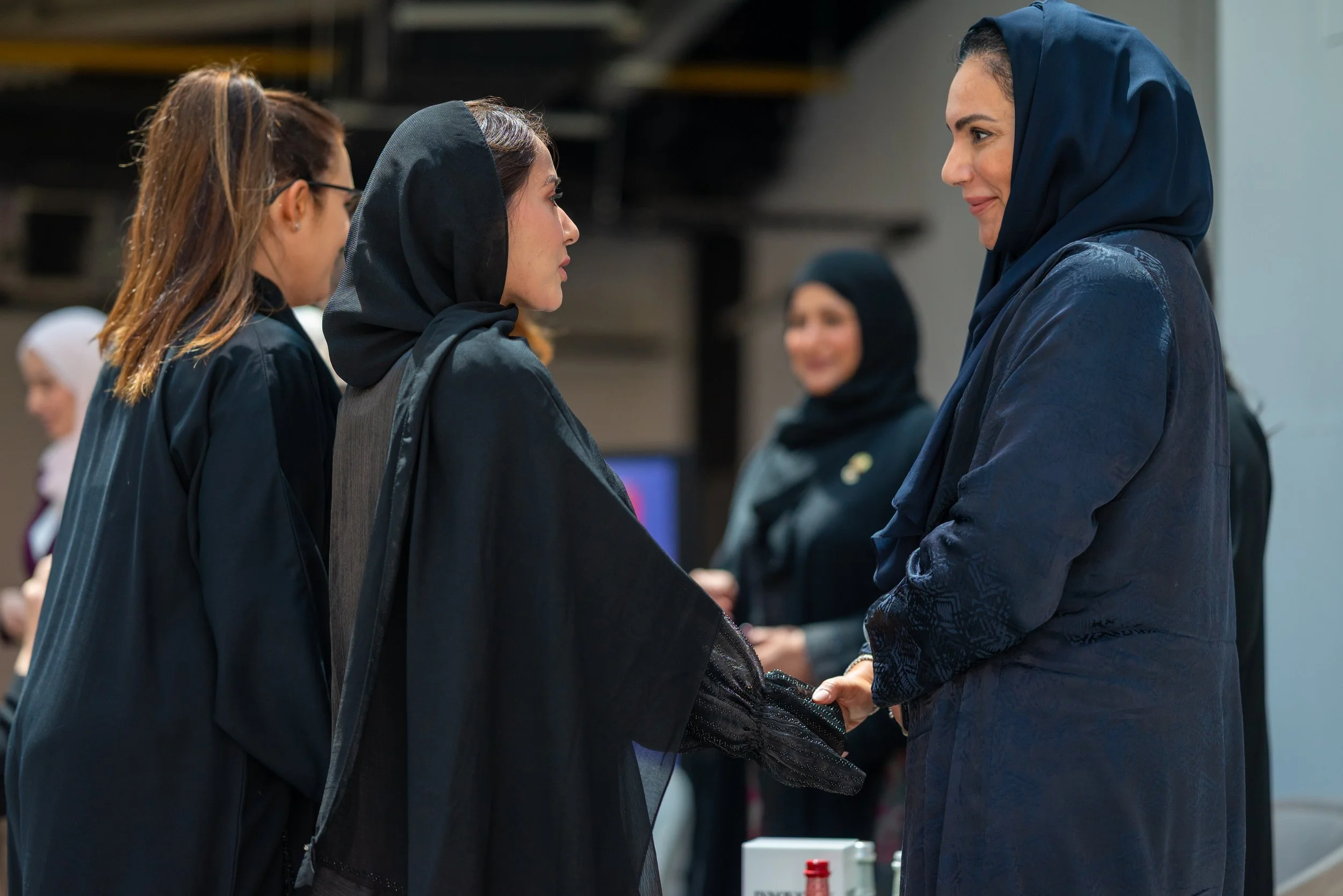 Women shaking hands at a formal event with others in the background.