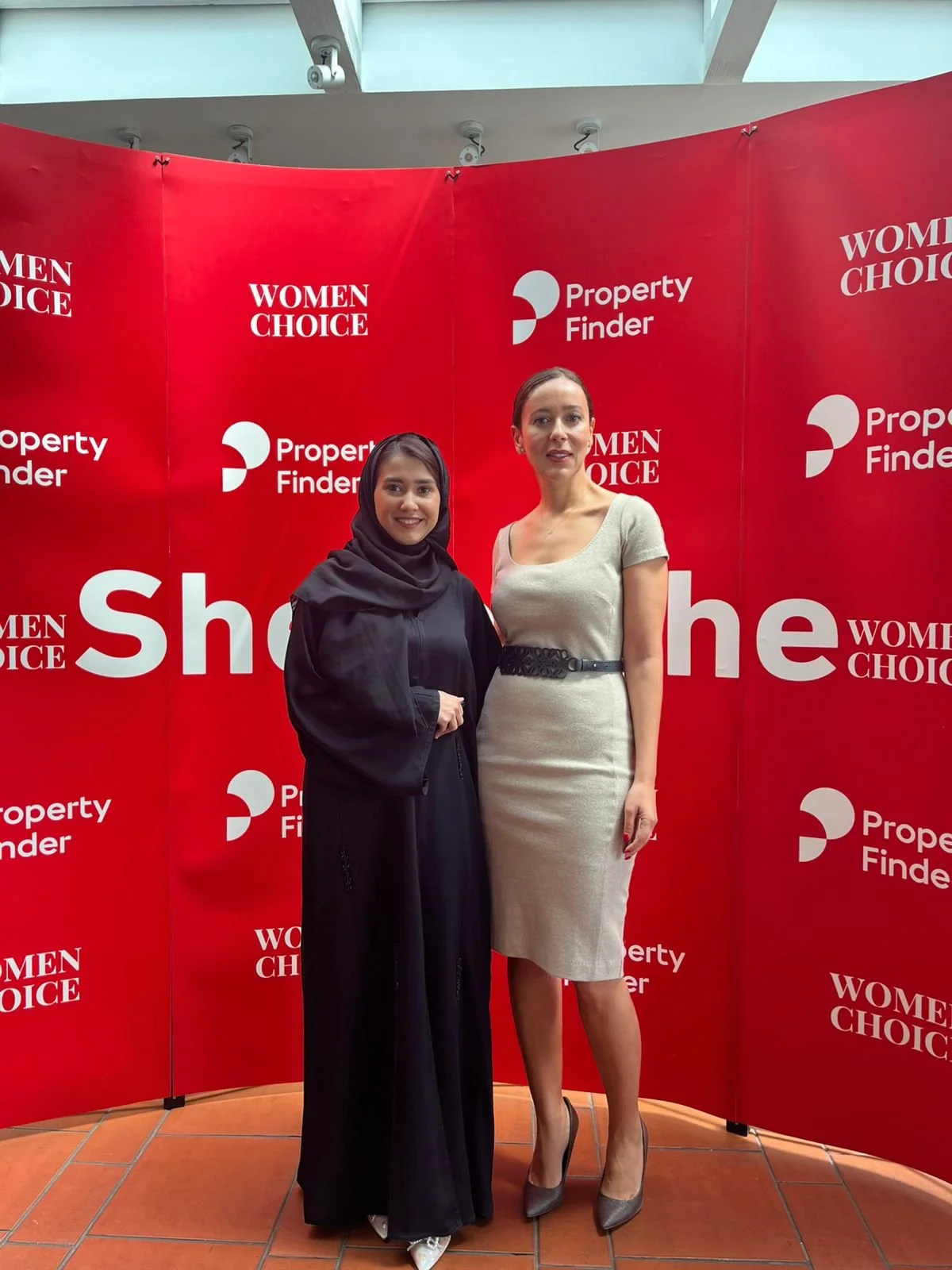 Two women standing side by side in front of a red backdrop with white text that reads "Women Choice," "Property Finder," and "Show." The woman on the left is wearing a black abaya and hijab, and the woman on the right is wearing a beige dress with a 