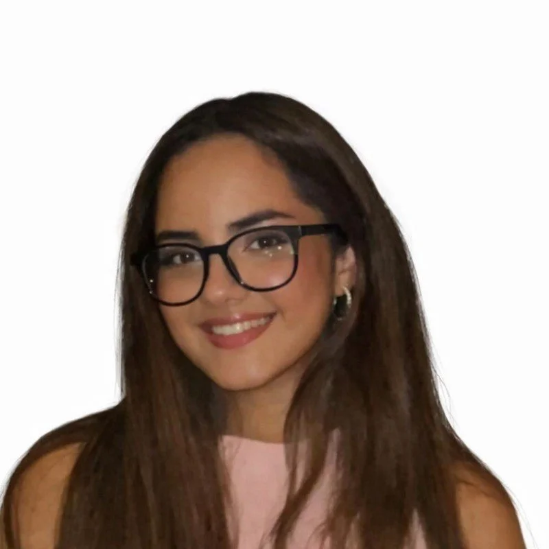 A smiling woman with shoulder-length black hair wearing a blue top in a lush greenhouse with green plants.