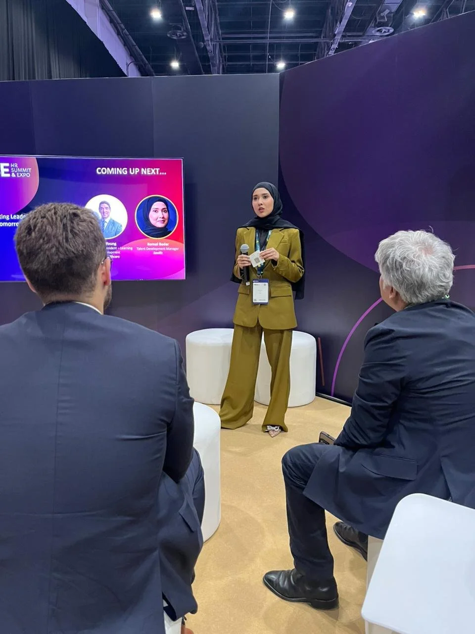 A woman in a mustard-colored suit and black hijab is giving a presentation at a conference, holding a microphone and standing near a digital screen that displays profiles of speakers, with two men in suits seated and listening attentively.