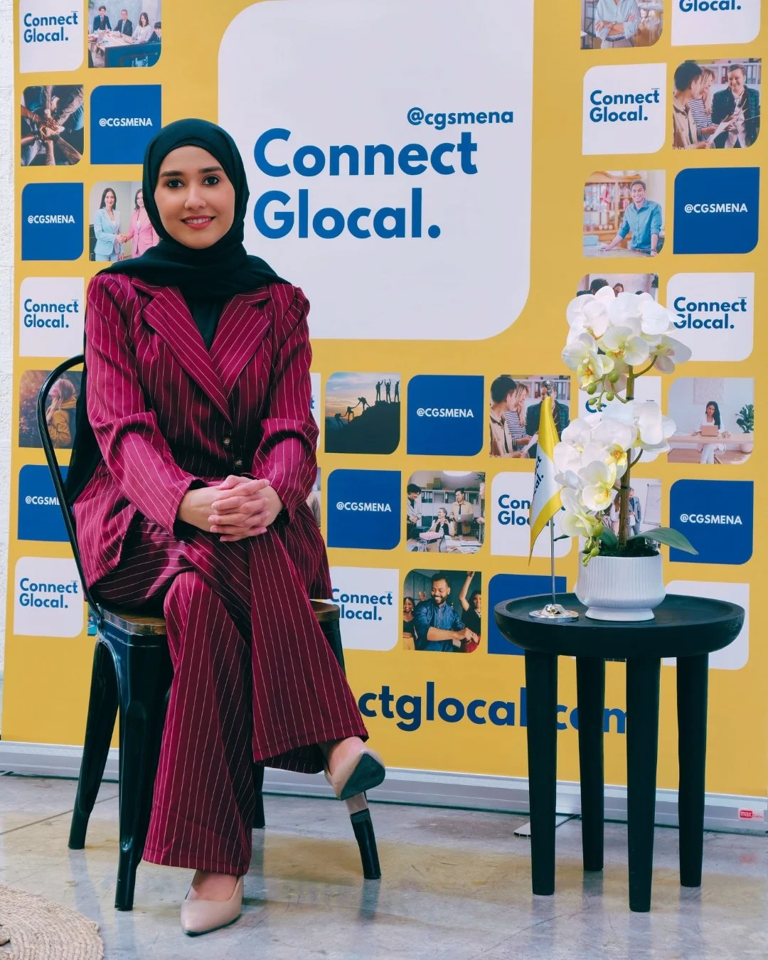 A woman wearing a maroon pinstripe suit and a black hijab, sitting on a black chair in front of a yellow backdrop with multiple social media logos and pictures. There is a small black table with a white orchid plant and a small flag to her right.