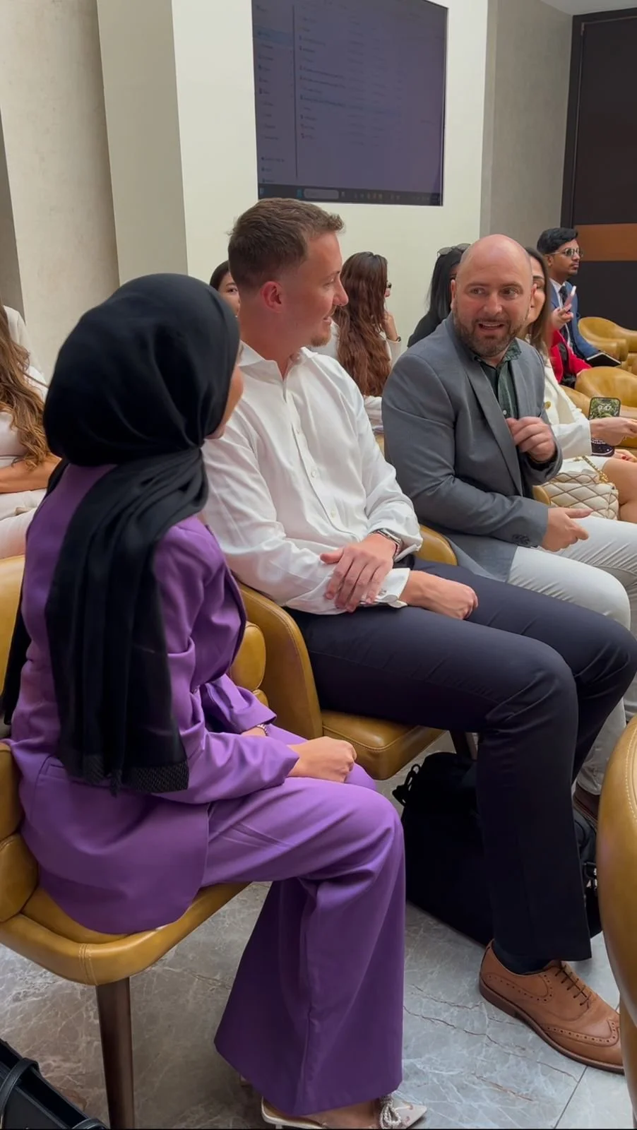 People sitting in a conference room, engaged in conversation. The woman in the foreground wears a purple suit and hijab, and the two men next to her are dressed in white and gray suits respectively.