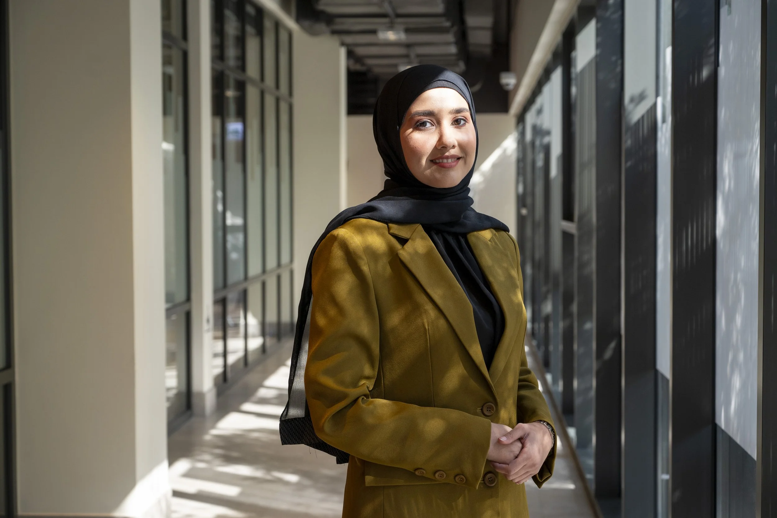A woman in a hijab standing in a modern building hallway with glass windows and sunlight.