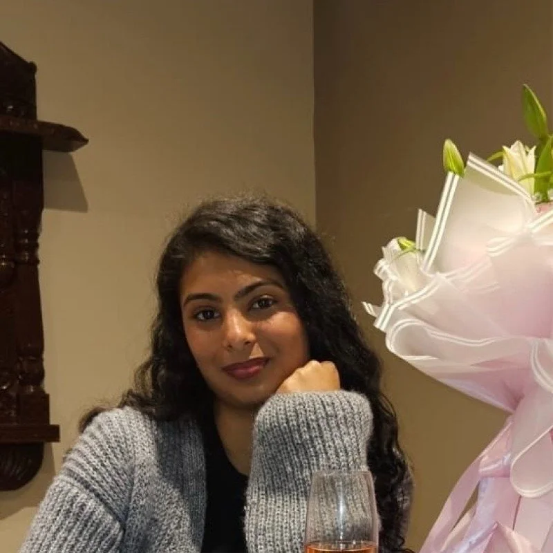 Smiling woman with curly hair wearing glasses and hoop earrings, in a room with blurred background.