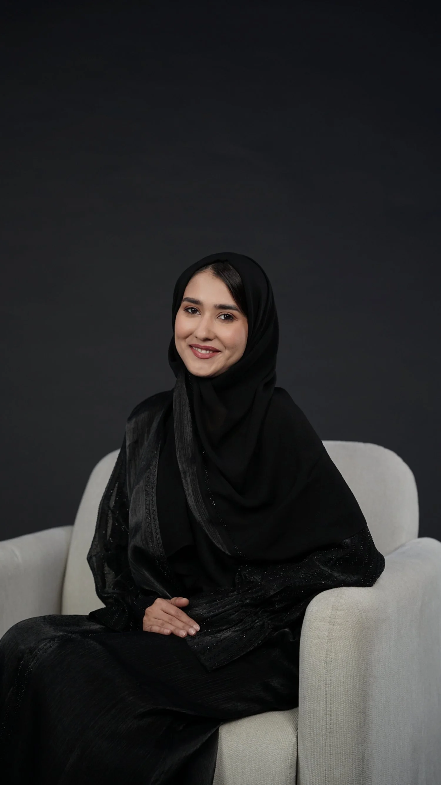 A woman wearing a black hijab and black outfit sitting on a light-colored armchair against a dark background, smiling at the camera.