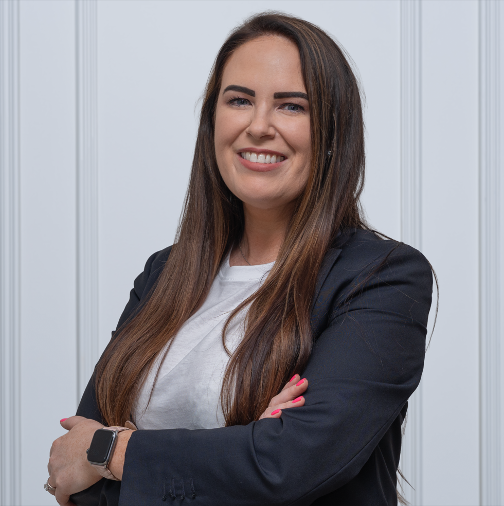 A woman with short black hair smiling in front of green plants in a greenhouse or botanical garden.