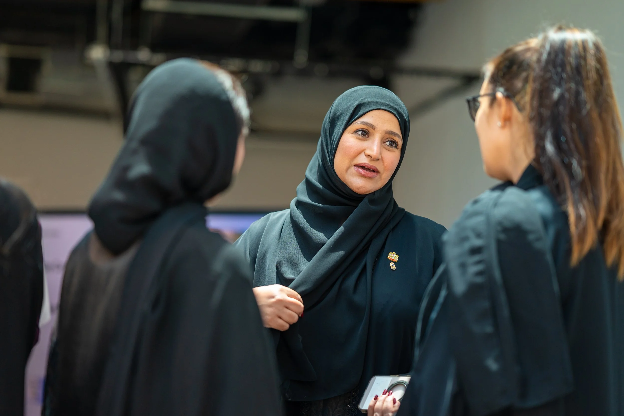 A woman wearing a hijab speaking to three women, one with a hijab and two wearing glasses, in an indoor setting.