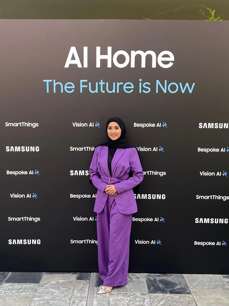 Woman in purple suit and black hijab standing in front of a Samsung and AI event backdrop with the text 'AI Home' and 'The Future is Now'.