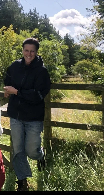 Woman smiling outdoors beside a wooden fence, surrounded by trees and greenery under a blue sky with clouds.