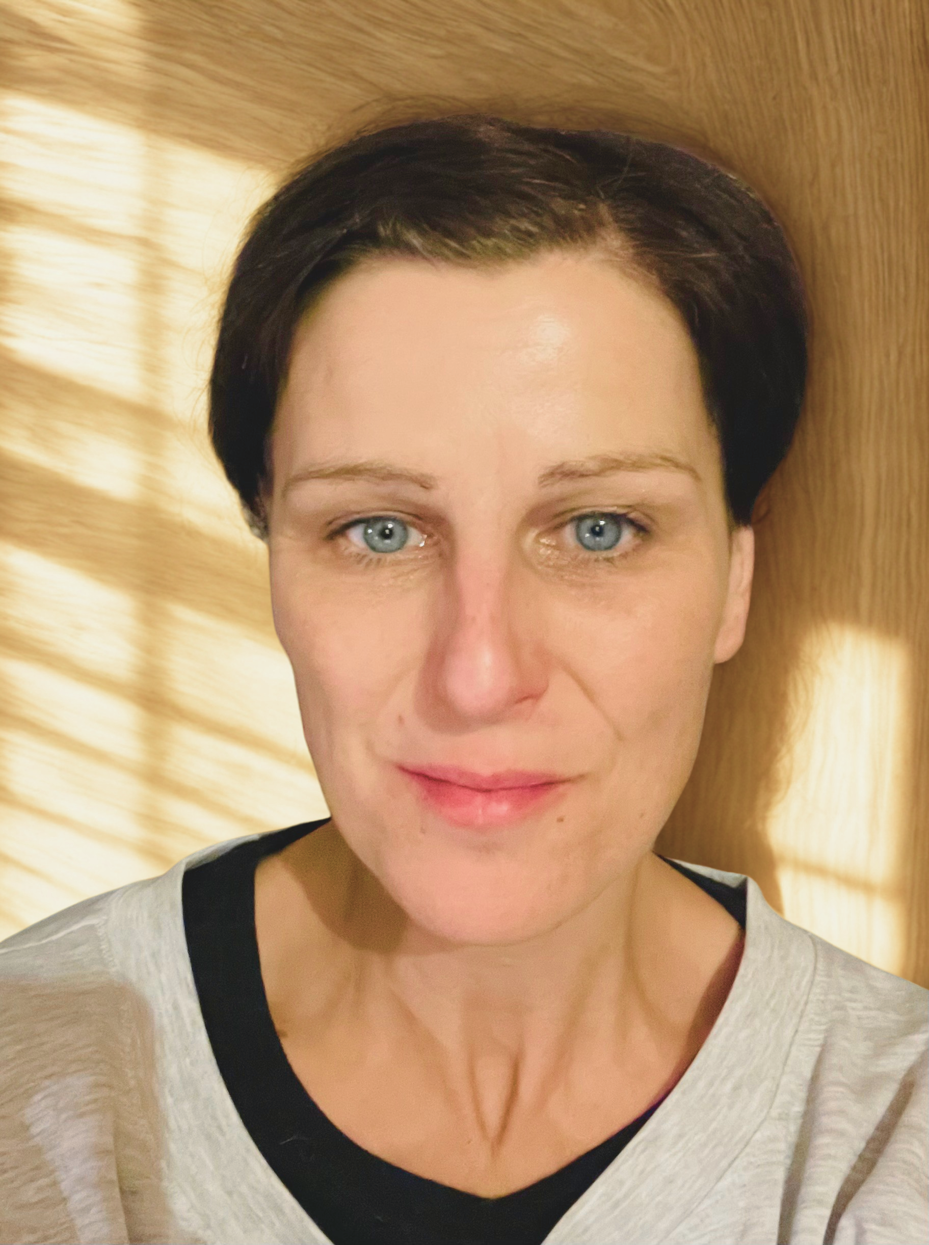 A woman with short dark hair and blue eyes is lying down on a wooden floor, looking directly at the camera with sunlight and shadows creating a pattern on the background.