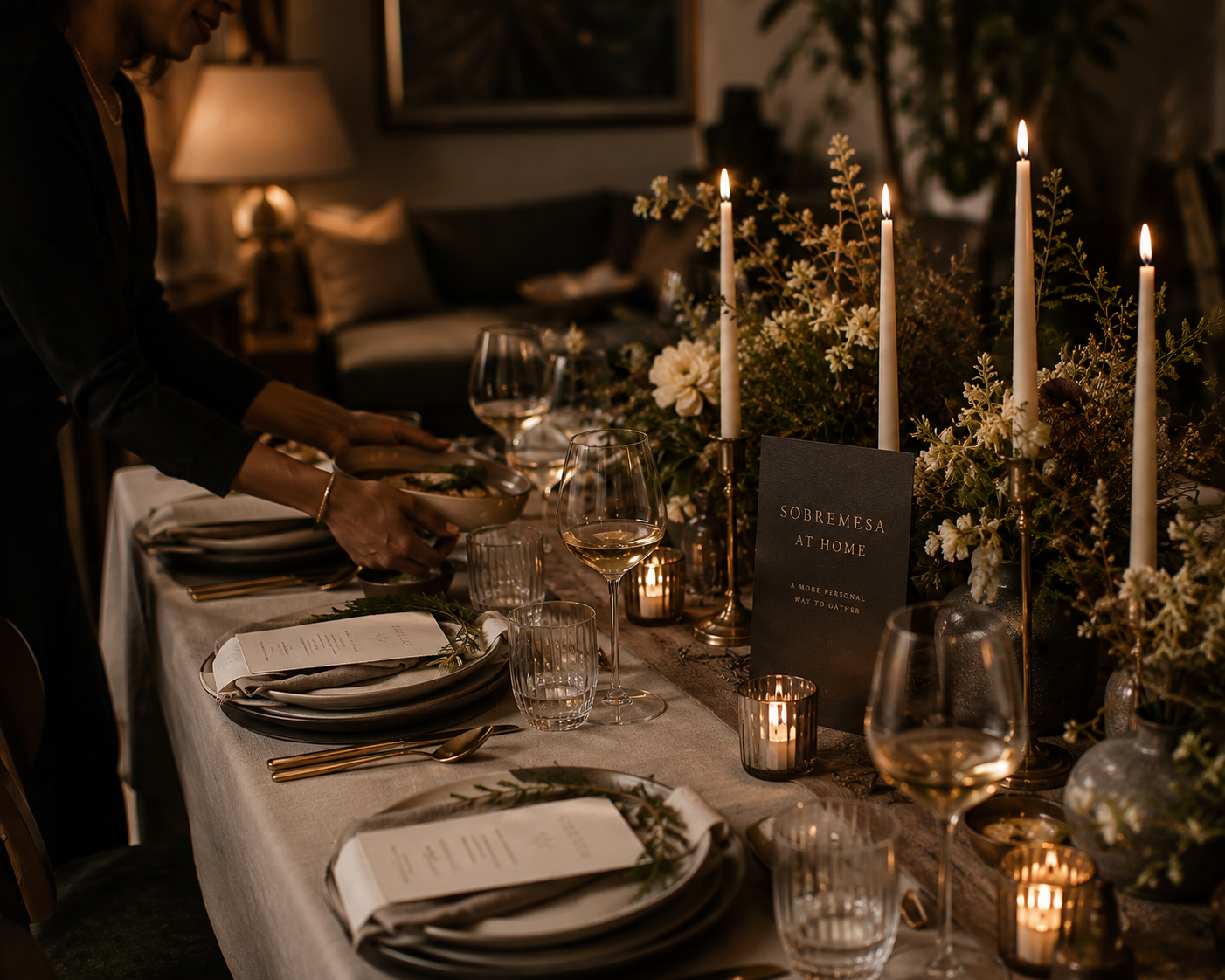 A dimly lit dinner table set with white plates, glassware, gold utensils, napkins, and a menu, decorated with tall candles, floral arrangements, and a sign that reads 'SOBREMESA AT HOME' in a cozy, elegantly decorated room.
