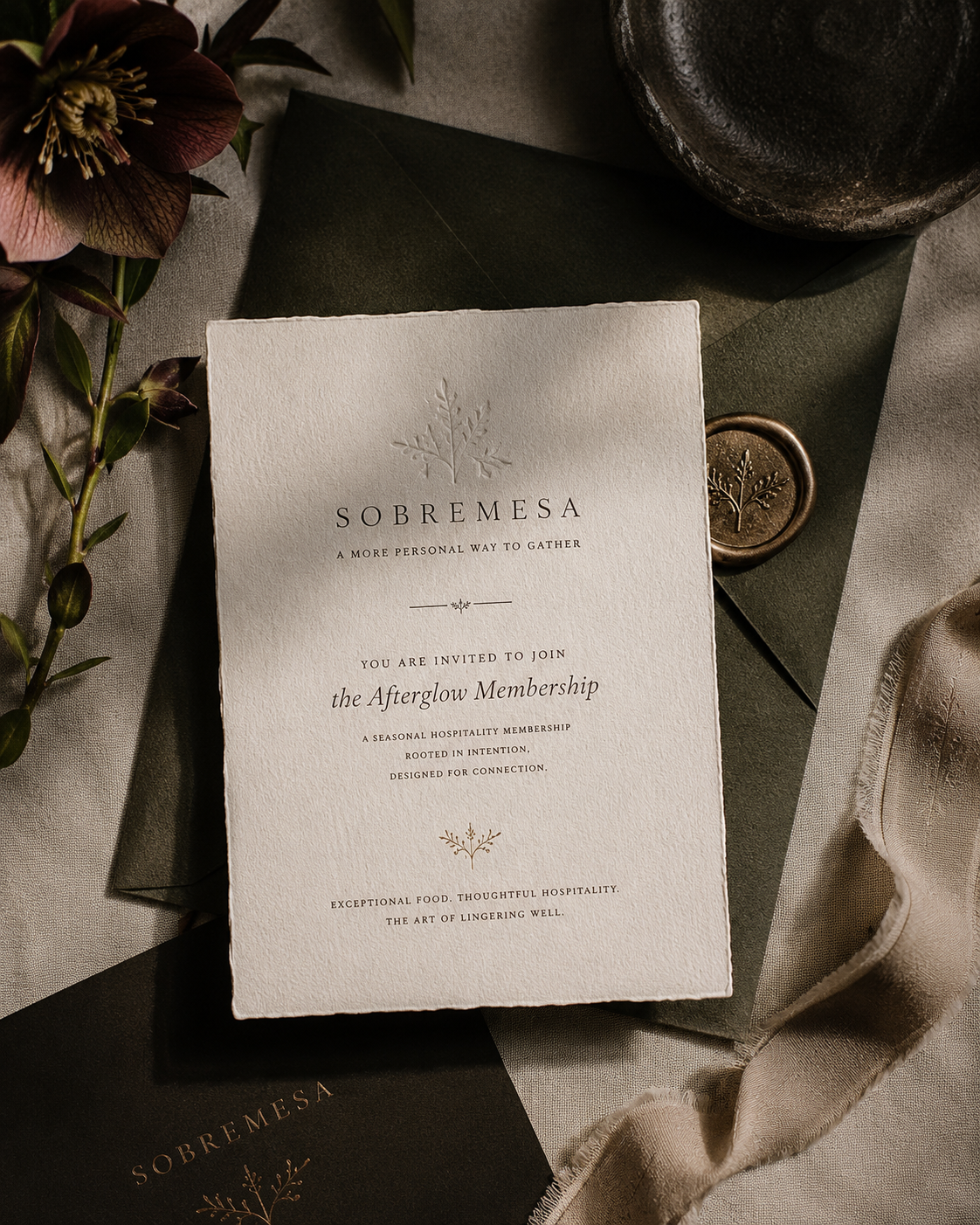 Invitation card for Sobremesa, a seasonal hospitality membership, surrounded by decorative items including a dark green envelope, a round metallic object, flowers, and ribbons.