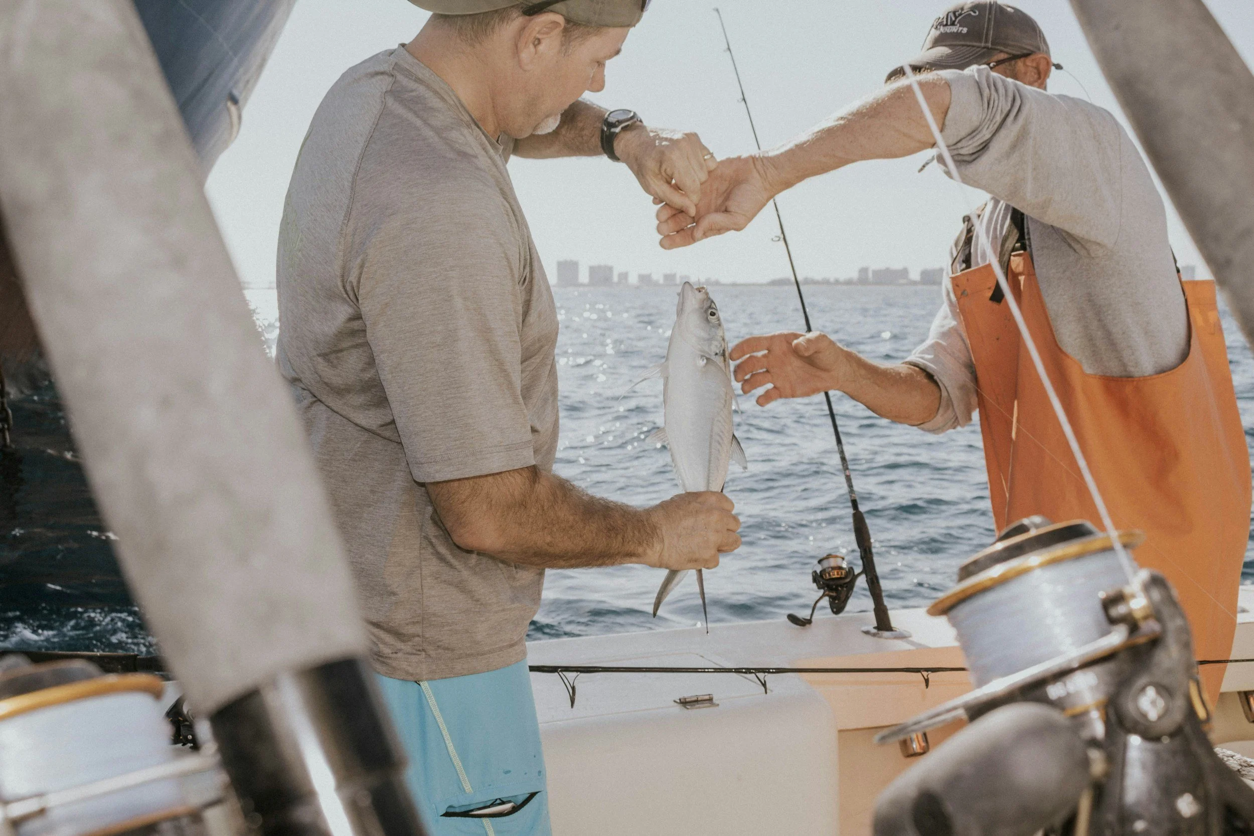 Two men on a boat, one holding a fish, the other reaching out, fishing gear visible, city skyline in the background.