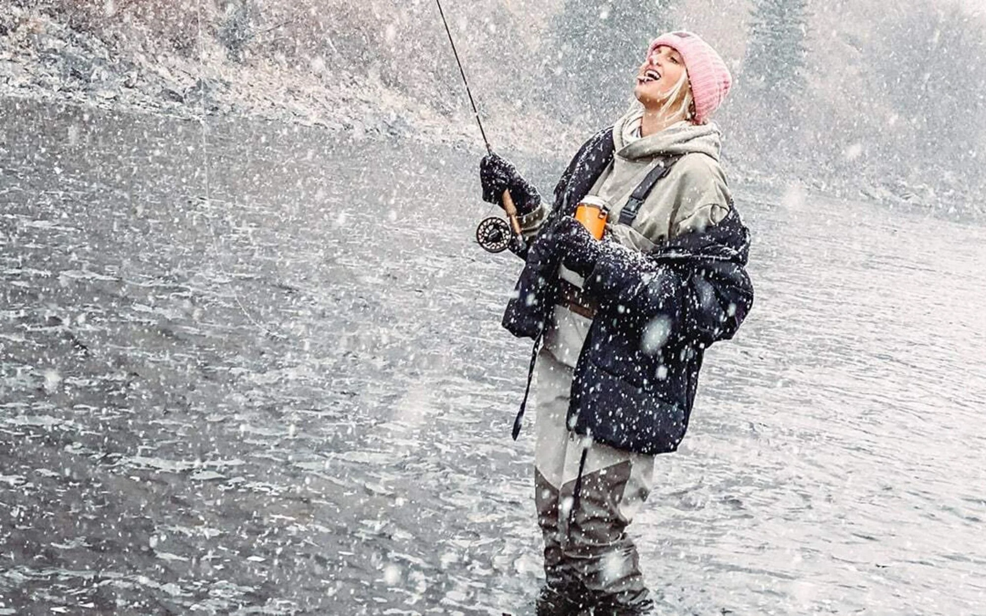 A woman fishing in a snowy lake, wearing a pink knit hat, black gloves, a beige and black winter jacket, and holding a fishing rod.