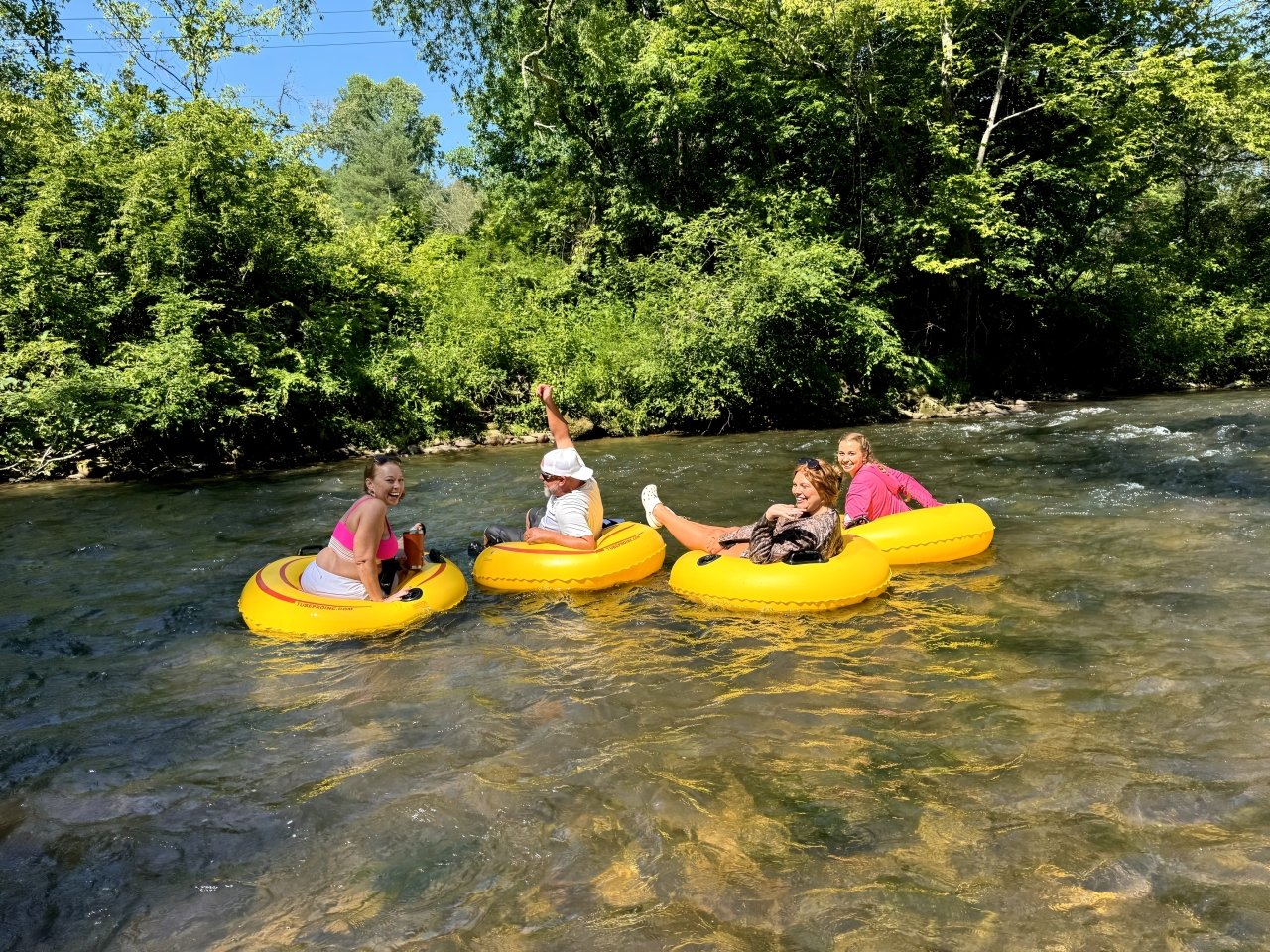 Four women on yellow inflatable tubes floating on a river surrounded by green trees, smiling and enjoying a sunny day.