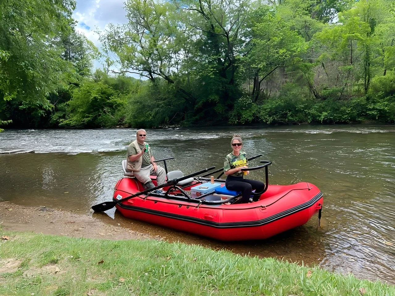 Two people sitting in a red inflatable boat on a river near a grassy bank, surrounded by lush green trees and foliage, with one person giving a peace sign and the other holding a drink, all wearing sunglasses.