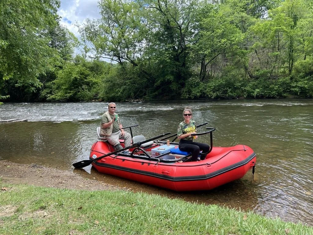 Two people in a red inflatable raft by a river with green trees in the background. One person is sitting on the edge of the raft holding an oar, and the other is sitting near some gear, holding a drink.