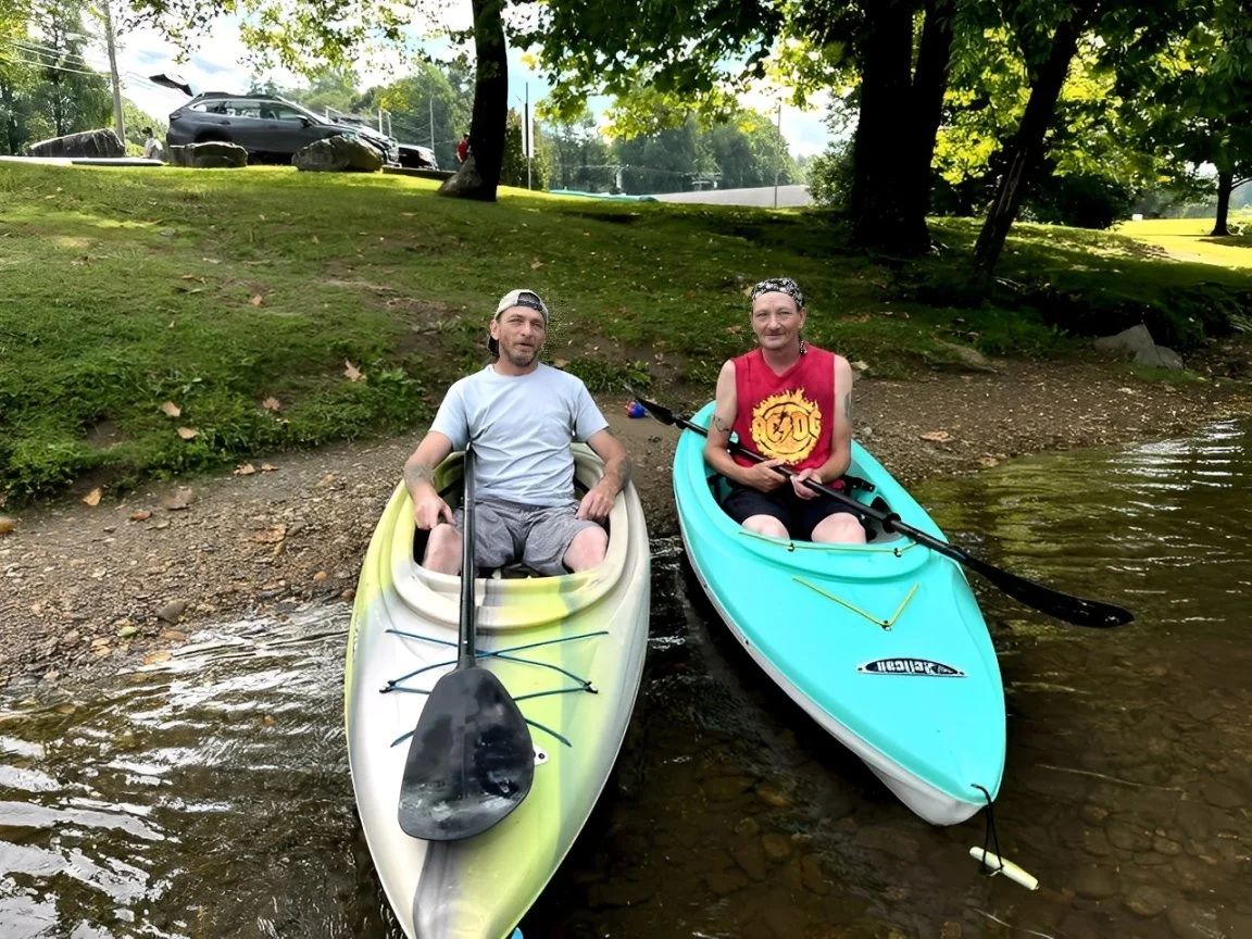 Two men kayaking on a small creek surrounded by trees and grassy banks.