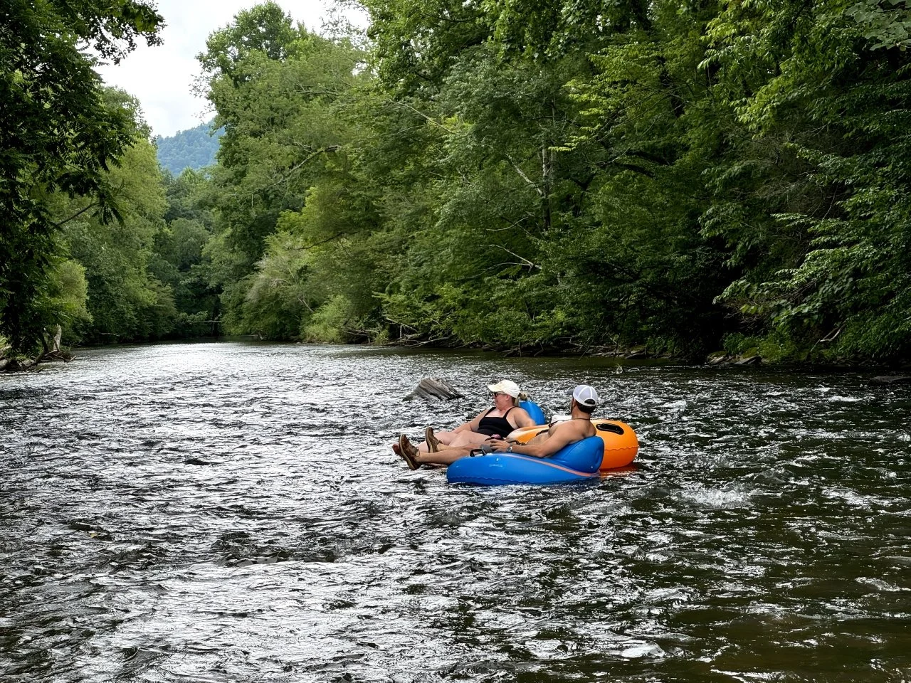 Two people floating on a river in inflatable kayaks surrounded by lush green trees.