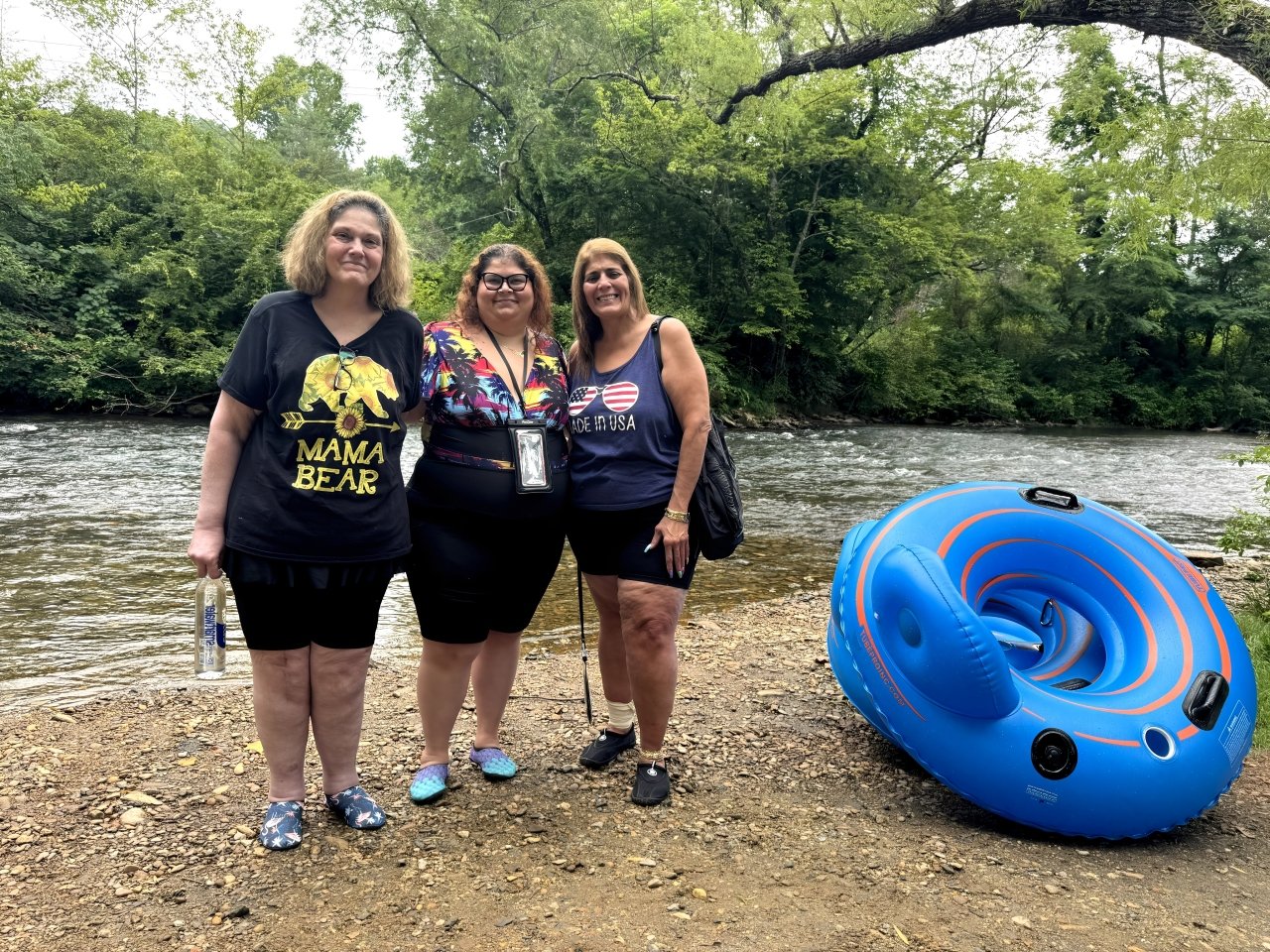 Three women standing by a riverbank with a blue inflatable raft nearby, surrounded by green trees.
