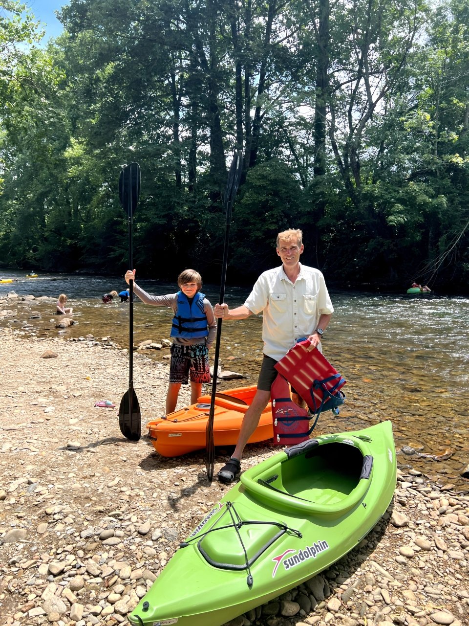 Two people, a child and an adult, standing by a river with kayaks and paddles, surrounded by trees, enjoying a summer day. The adult is holding a red life vest, and the child is wearing a blue life vest.