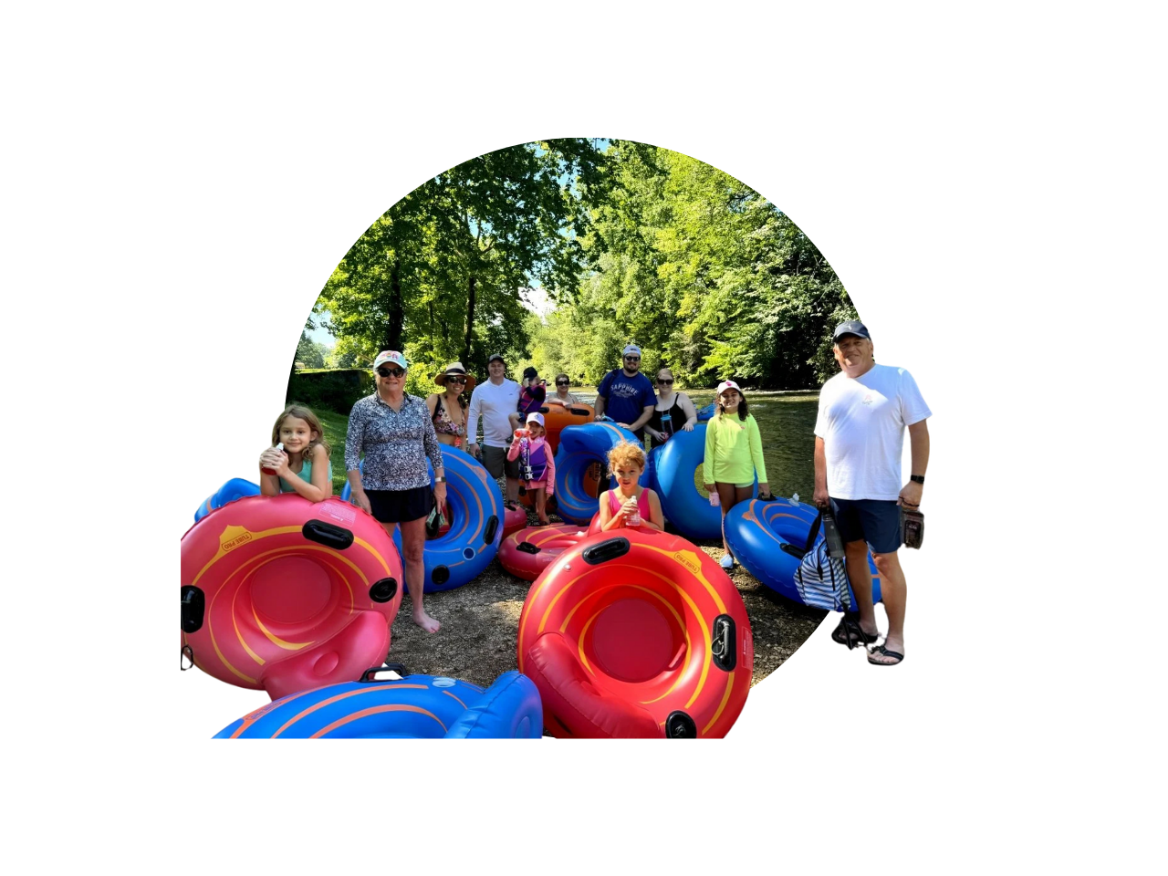 Group of people, including children, in a wooded outdoor area by a river, holding colorful inflatable tubes for tubing, on a sunny day.