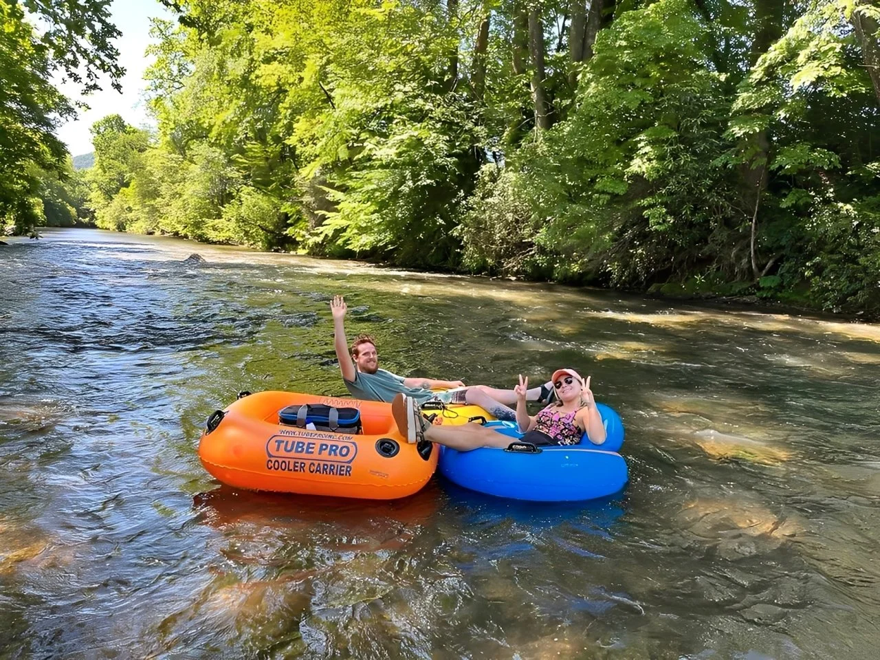 Two people floating on a river in inflatable tubes surrounded by green trees, one in an orange tube and the other in a blue tube, smiling and making peace signs.