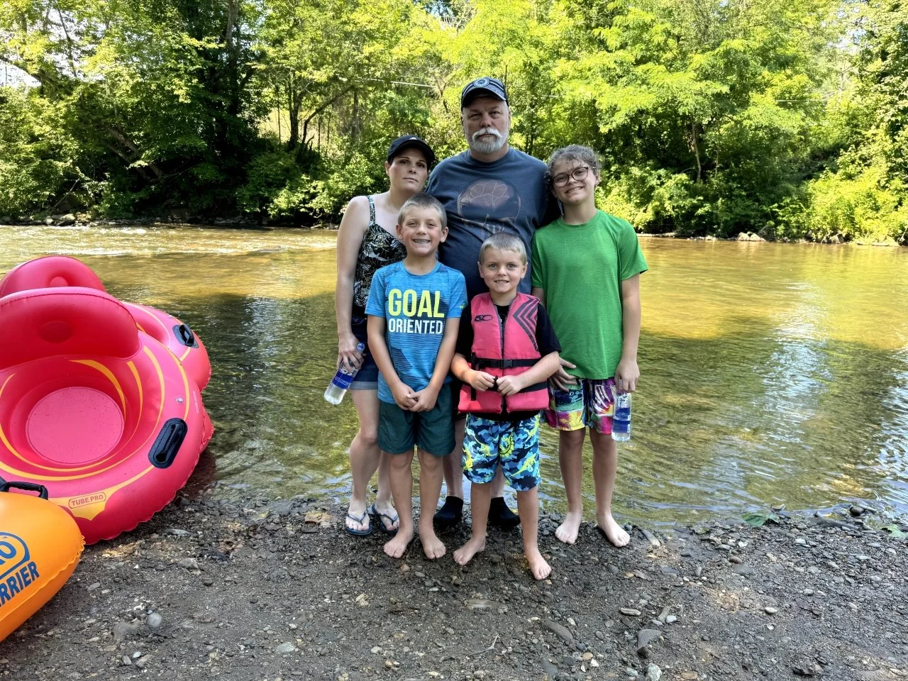 A family of six standing on a riverbank with trees in the background, wearing swimsuits and life jackets, with inflatable tubes nearby.
