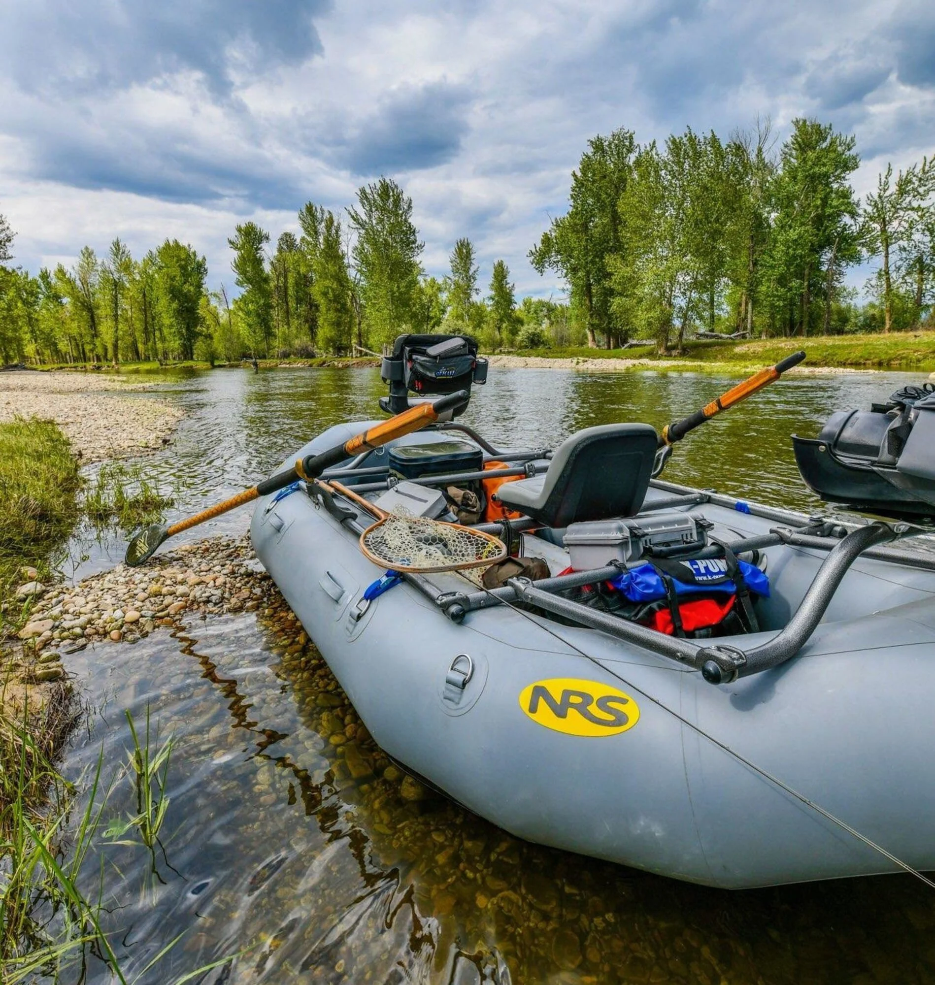 An inflatable gray boat with fishing gear, paddles, and a seat, resting on a pebbled riverbank with lush green trees and cloudy sky in the background.