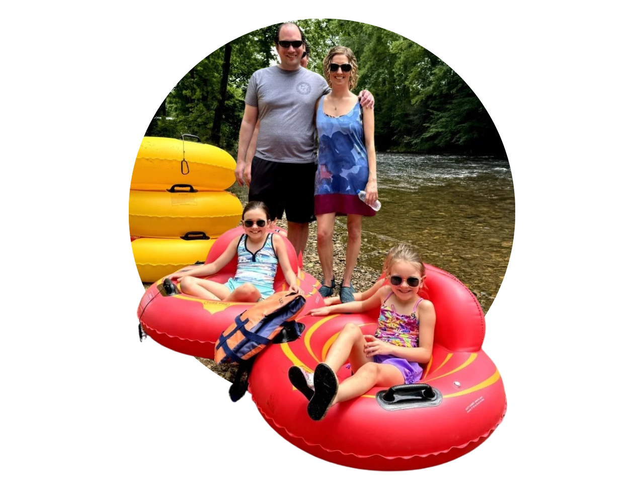A family of four at a river, with two women standing and two young girls sitting on a red inflatable raft, beside yellow tubes, surrounded by green trees.