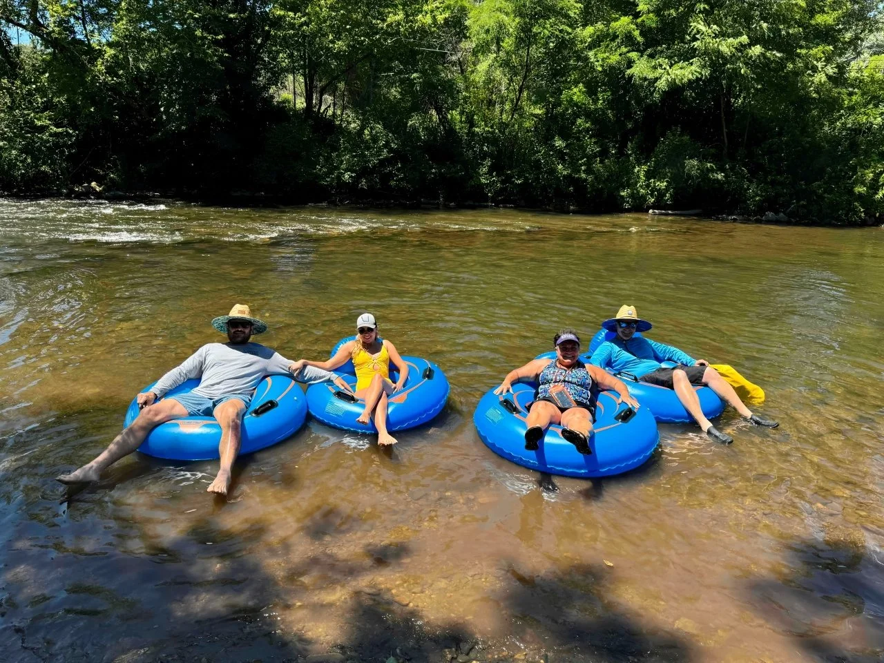 Four people relaxing in blue inner tubes in a shallow river, surrounded by greenery, sunny weather, wearing casual summer clothes and hats.