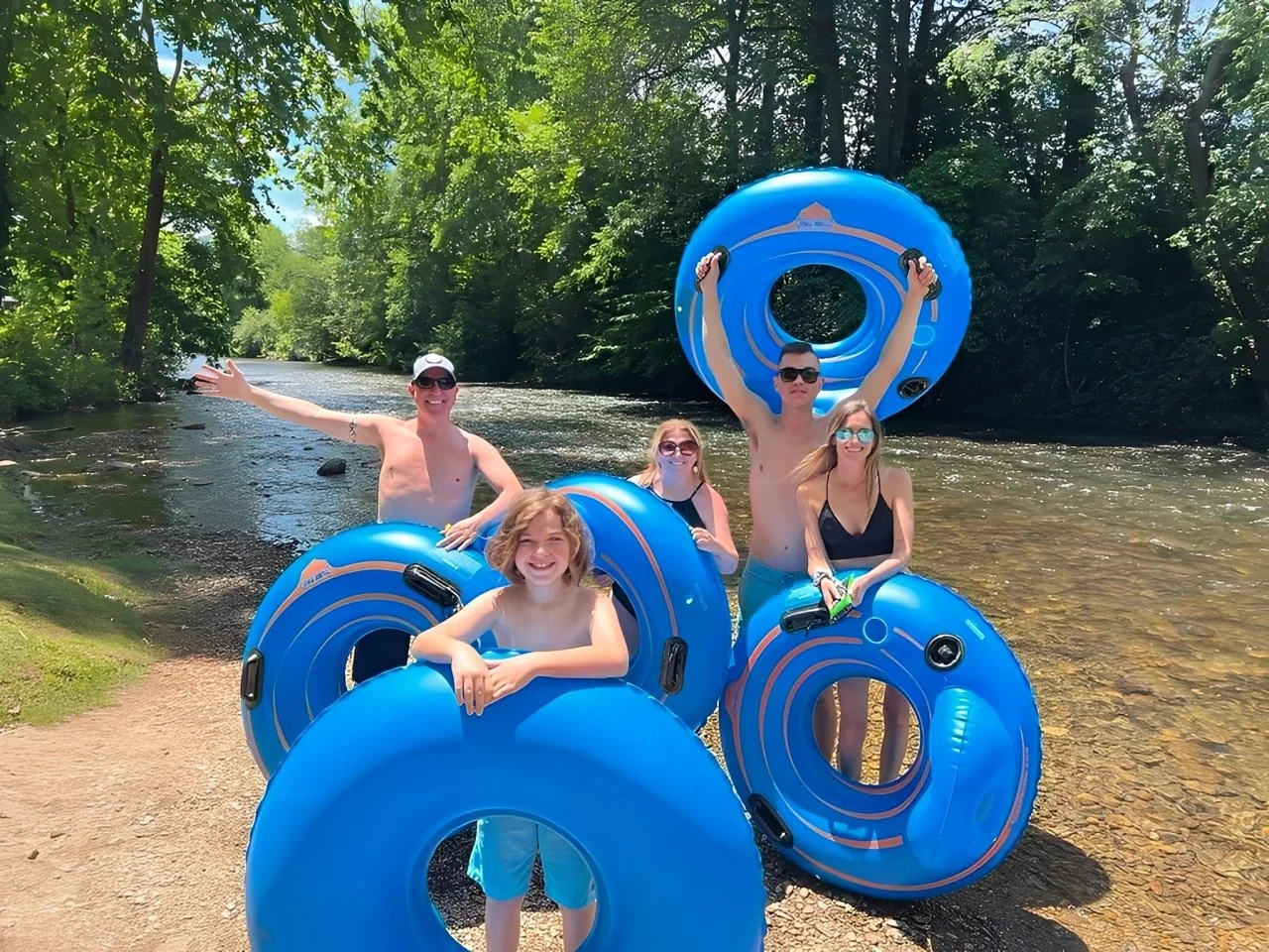 Group of five people in blue inner tubes on a river, surrounded by lush green trees, enjoying a summer day.