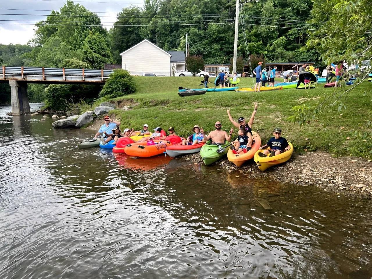 People kayaking and preparing to kayak along a lake shoreline, with some sitting in kayaks near the water and others on the grassy bank, surrounded by trees and a bridge in the background.