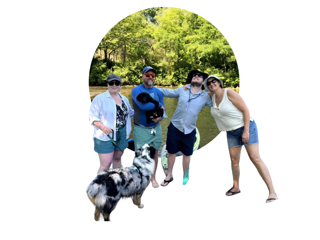 A group of four people and two dogs outdoors by a river, smiling and enjoying a sunny day.