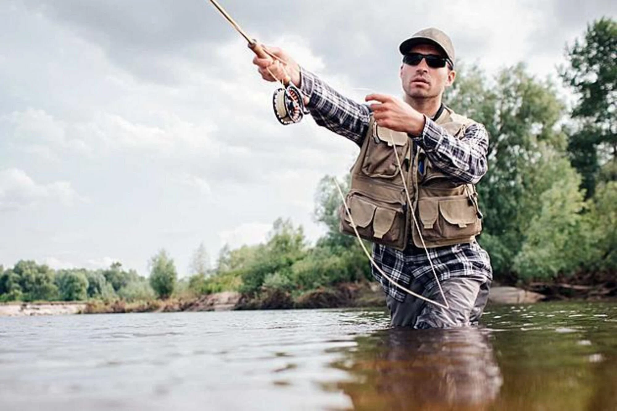 A man wearing sunglasses, a cap, a fishing vest, and plaid shirt stands in a river, fishing with a rod and reel, with trees and cloudy sky in the background.